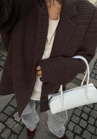 Dunkelbrauner, gestrickter oversized Blazer mit einem strukturierten Muster, weißes Oberteil, graue, strukturierte Hose und eine weiße rechteckige Handtasche.
