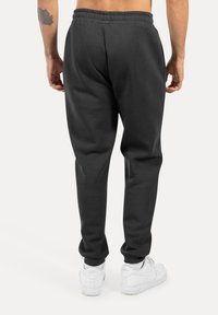 Černé tepláky vyrobené z měkké tkaniny, s elastickým pásem a manžetami, v kombinaci s bílými teniskami. Pořízeno ze zadu.