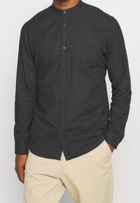 Hombre vestido con una camisa de manga larga gris oscuro con cuello mao y pantalones beiges, con las manos en los bolsillos, frente a un fondo liso.