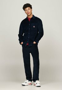 Chemise en velours côtelé bleu marine et pantalon assorti, avec une poche à rabat, un tissu texturé et des baskets blanches avec des accents rouges et bleus.