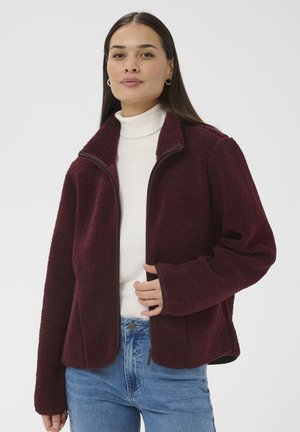 Frau trägt eine bordeauxrote Fleecejacke über einem weißen Rollkragenpullover und blauen Jeans, posiert mit einer Hand am Reißverschluss der Jacke.