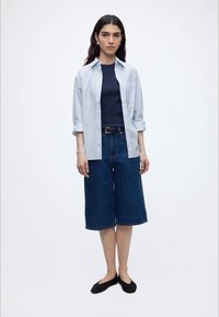 Nainen seisoo, hänellä on päällä vaaleansininen napitettuna oleva paita laivastonsinisen topin päällä, tummat farkkukneepparit, mustat ballerinat ja helmirenkaat valkoista taustaa vasten.