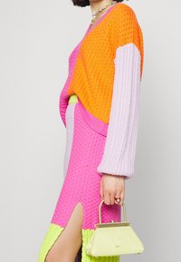 Kolorowy dziany sweter w odcieniach pomarańczu, różu i lawendy; dopasowana spódnica z bocznym rozcięciem i jasnymi zielonymi akcentami. Trzyma żółtą torebkę ze złotymi detalami.