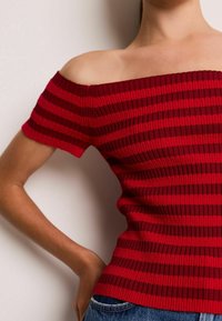 Red and dark red striped ribbed off-shoulder top with short sleeves, fitted design, and textured fabric. Worn with blue denim jeans.