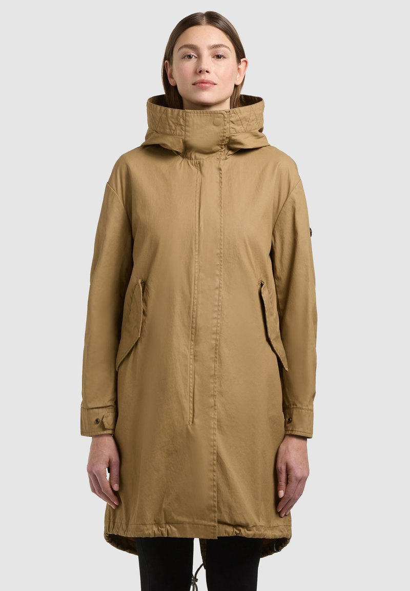 Kobieta ubrana w beżowy, sięgający kolan płaszcz z kapturem, z kieszeniami z klapkami i zapinanymi na guziki mankietami, stojąca na tle jednolitego jasnego tła.