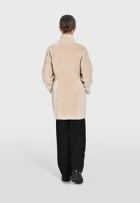 Abrigo de peluche sintético beige claro con cuello, mangas largas y corte recto, combinado con pantalones de pierna ancha negros y zapatos negros gruesos.