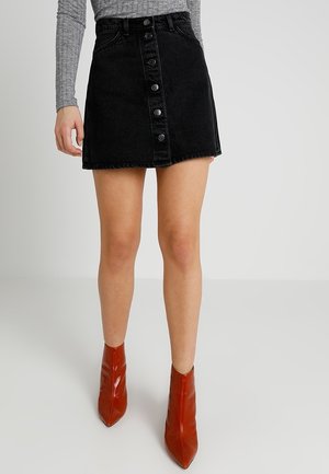 Kvinde iført en sort denim mininederdel med knapper foran, grå langærmet top og spidse røde ankelstøvler, stående på en neutral baggrund.