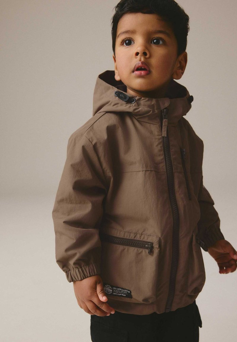 Junger Junge trägt eine braune Kapuzenjacke mit Reißverschluss und elastischen Bündchen und schaut leicht nach oben vor einem schlichten Hintergrund.