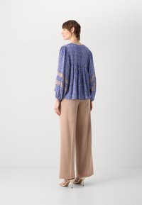Blå mønstret bluse med lange, puffede ærmer, en samlede ryg, parret med beige brede bukser og strop sandaler.