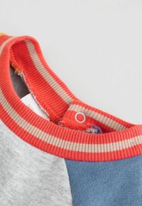 Mehrfarbiger T-Shirt-Kragen mit rotem Rippenbund, der beige Streifen aufweist; enthält einen Druckknopfverschluss und verschiedene strukturierte Stoffeinsätze in Grau und Blau.