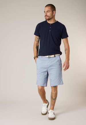 Homme tatoué portant un t-shirt henley bleu marine, un short bleu clair avec une ceinture blanche, et des baskets blanches, marchant sur un fond uni.