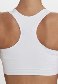 Hvid sports-bh med racerback-design, der har et tekstureret stof og en ribbet kant forneden. Bagsidevisning, ingen synlig hardware.