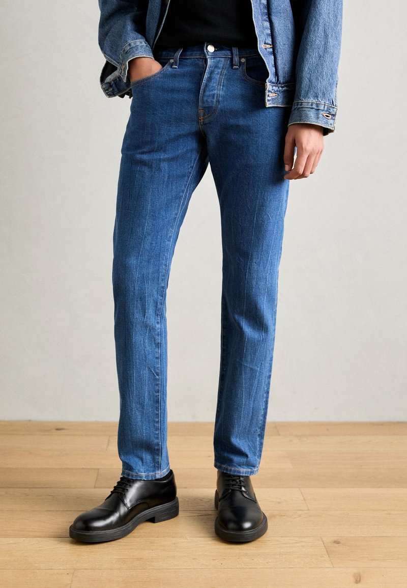 Blå denimjeans med ljus blekning, åtsittande skärning, femfickorsdesign och medelhög midja, tillsammans med svarta läderskor.