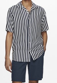 Uomo che indossa una camicia a maniche corte a righe verticali blu e bianche con pantaloni corti texturizzati blu, con una mano in tasca.