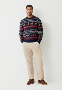 Mann steht mit Händen in den Taschen, trägt einen festlichen Pullover mit Hundemuster, beige Cordhosen und braune Lederschuhe vor einer weißen Wand.