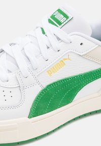 Biały skórzany tenisówka z zielonym zamszowym akcentem. Posiada złote logo, białe sznurowadła oraz teksturowaną kremową gumową podeszwę.