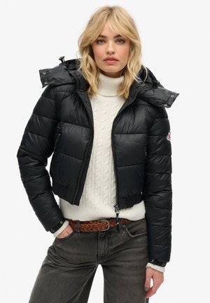 Schwarze Daunenjacke mit cropped Design, mit Kapuze, Reißverschluss und horizontalen Steppnähten. Getragen über einem cremefarbenen Strickpullover.