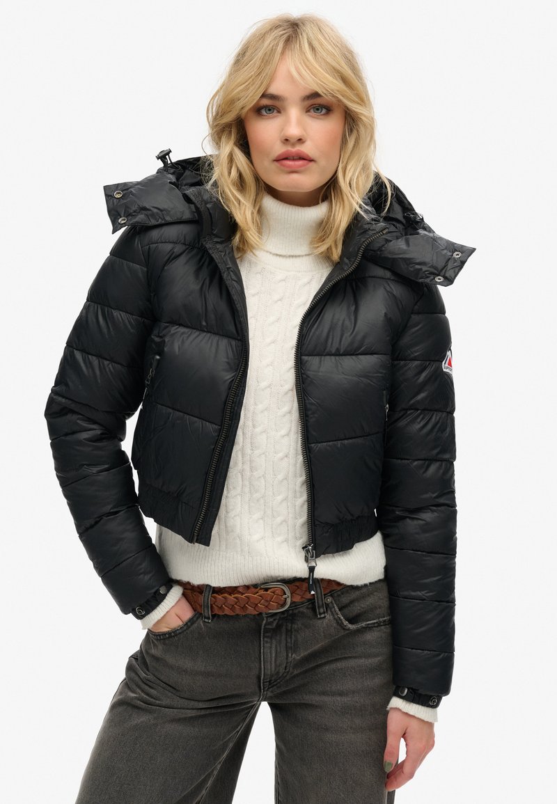 Giacca a vento nera con design corto, dotata di cappuccio, chiusura con zip e trapuntatura orizzontale. Indossata sopra un maglione lavorato a maglia color crema.