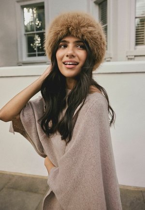 Round, fluffy tan fur hat; soft, beige knit poncho with wide sleeves; smooth, dark hair; light background with window framing.