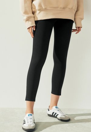 Zwarte leggings van rekbaar materiaal met een slank ontwerp, gecombineerd met wit- en grijsgekleurde sneakers met zwarte strepen en blauwe accenten.