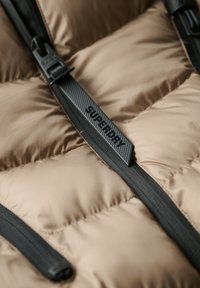 Beige pufferjakke stof med en glat, skinnende overflade, udstyret med sorte lynlåse og et vævet mærke med teksten "SUPERDRY" på lynlåstrækkeren.