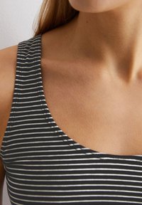 Gestreiftes ärmelloses Oberteil in Schwarz und Weiß mit einem runden Ausschnitt und breiten Trägern, hergestellt aus weichem, strukturiertem Stoff. Nahaufnahme von Schulter und Schlüsselbein.