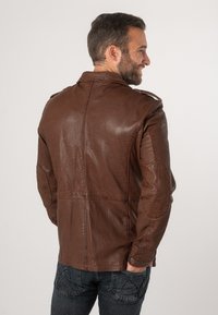 Braune Lederjacke mit strukturiertem Finish, Schulterepauletten und Nahtdetails. Über dunklen Jeans getragen, zeigt einen lässigen Schnitt.