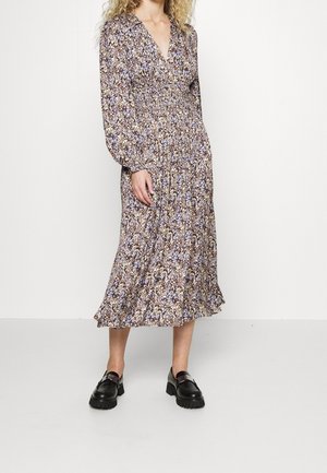 Frau trägt ein langärmliges, florales Midikleid mit V-Ausschnitt und schwarzen, klobigen Loafers, steht vor einem schlichten weißen Hintergrund.