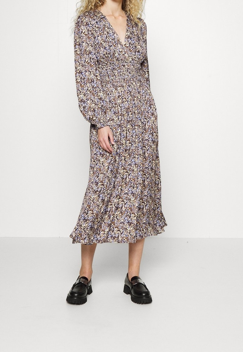 Femme portant une robe midi à manches longues avec un motif floral, un col en V et des mocassins noirs épais, debout devant un fond blanc uni.