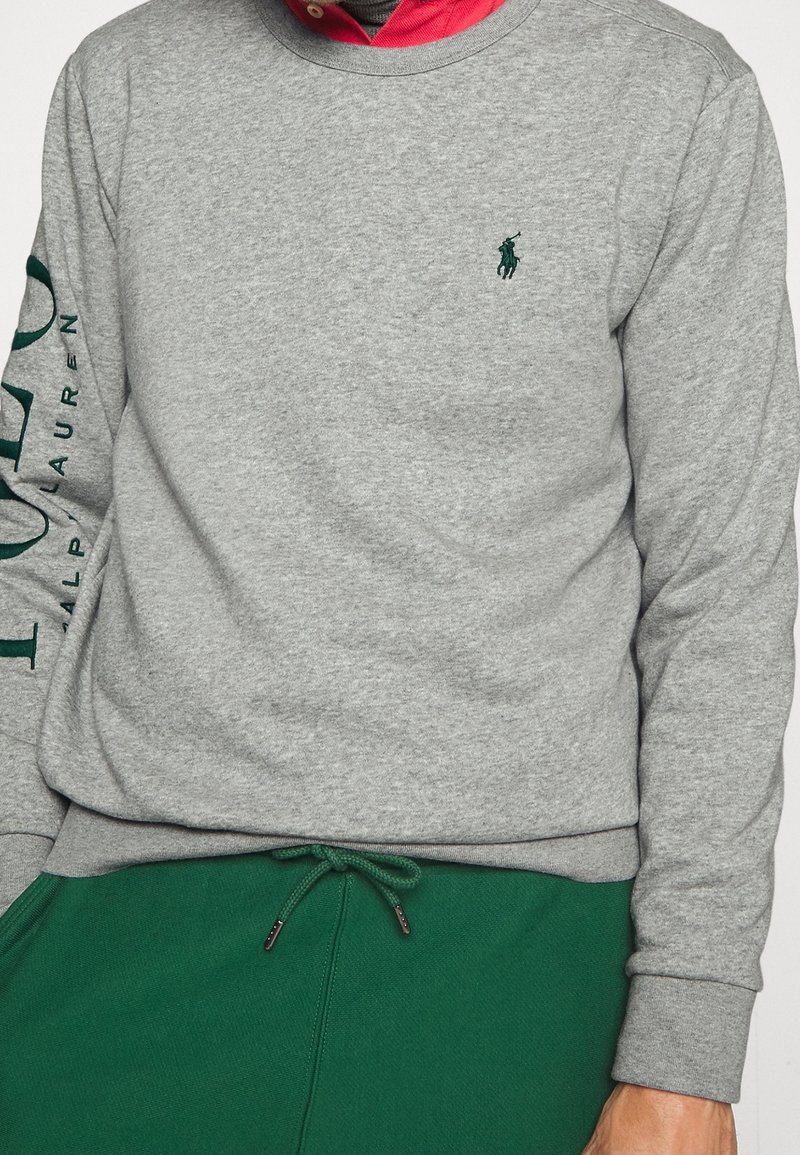 Chemise à manches longues grise avec logo Ralph Lauren vert sur la poitrine et la manche, portée sur une chemise rouge à col, assortie à un pantalon vert à cordon de serrage.