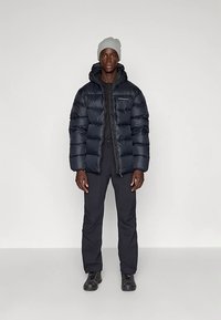 Navy puffer jacket with a hood, featuring horizontal stitching and a front zipper. Paired with black pants and a light gray hat.
