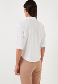 White short-sleeve shirt with a textured finish and a curved hem, paired with beige pants. The shirt features a relaxed fit and a collar.