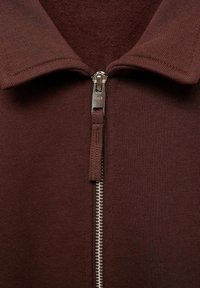 Burgundská zipová bunda s límcem, žebrovanou texturou a stříbrným zipem. Detailní pohled zdůrazňuje látku a zpracování.