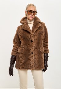 Brauner Kunstfellmantel mit Schlangenmuster, zwei vorderen Taschen, Reverskragen und schwarzen Knöpfen. Ergänzt durch lange braune Lederhandschuhe.