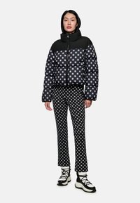 Schwarze Daunenjacke mit weißem Sternenmuster, hohem Kragen und Reißverschluss, kombiniert mit passenden Sternenmuster-Hosen und schwarzen Schnürstiefeln.