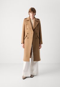 Palton din lâna de cămilă, cu două rânduri de nasturi, guler ascuțit, nasturi aurii, buzunare laterale și un design drept. Purtați peste un top bej și pantaloni deschiși.