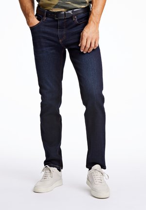 Jeans de mezclilla azul oscuro con costuras naranjas, cinco bolsillos y corte slim, combinados con zapatillas blancas.