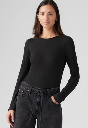 Langärmliges, enganliegendes schwarzes Oberteil aus weichem Material. Kombiniert mit hochgeschnittenen dunklen Jeans mit Kordelzug und vorderen Taschen.