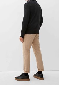 Schwarzes Langarmshirt, beige Chinos und schwarze Sneaker mit brauner Sohle. Die Schuhe haben eine glatte Textur; die Hose ist an den Saum umgeschlagen.
