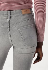 Grijze denim jeans met een hoge taille, met een tan leren label en achterzakken met subtiele stiksels. Gestreepte top zichtbaar bij de taille.