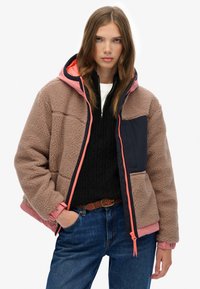 Fleece-lined hooded jacket in beige with a navy panel and pink accents. Features a front zipper and a chest pocket. Casual fit with a textured design.