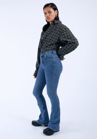 Camisa de cuadros oversized en negro y verde, vaqueros acampanados de cintura alta en azul y zuecos negros. Tejido suave y diseño estructurado.