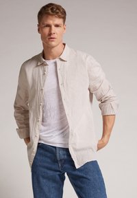 Camisa de lino color beige claro con botones en la parte delantera y mangas largas. Llevada sobre una camiseta blanca y combinada con unos vaqueros azules. Tejido simple y texturizado.