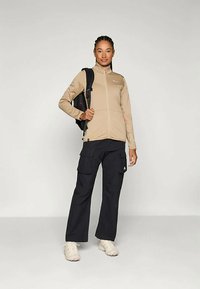Beige Zip-Up-Jacke mit hohem Kragen, schwarze Cargo-Hosen und einen schwarzen Rucksack. Helle Schuhe vervollständigen das Outfit.