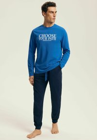 Man standing barefoot wearing blue long-sleeve shirt with "CHOOSE YOUR PATH" text and dark blue jogger pants against plain background.