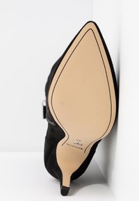 Chaussure à talon haut noire à bout pointu avec un extérieur en daim, présentant une semelle beige et un talon étroit. Détails du logo et de la taille visibles sur la semelle.