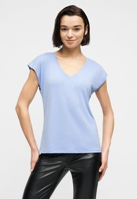 Νέα γυναίκα με κοντά σκούρα μαλλιά, που φοράει ανοιχτό μπλε μπλουζάκι με V και κοντά μανίκια, καθώς και μαύρο δερμάτινο παντελόνι, στεκόμενη με τα χέρια στη μέση.