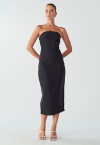 Žena v černých midi šatech bez ramínek a bílých sandálech na vysokém podpatku, stojící s rukama za zády na jednobarevném světlém pozadí.