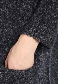 Czarny sweter o fakturowanym wykończeniu. Posiada podwinięty rękaw i boczną kieszeń, prezentując mieszankę szarych włókien.