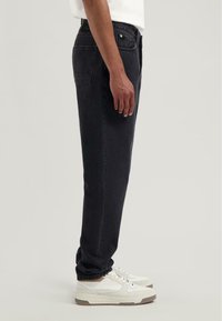 Czarne dżinsy o prostym kroju, z dwiema tylnymi kieszeniami i paskiem wyposażonym w zieloną metkę marki. Noszone z białymi trampkami.